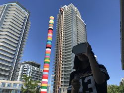 Image of a woman holding a virtual reality headset with her left hand. Behind her are two tall condominiums and between them stands a tall, multicolour poll.