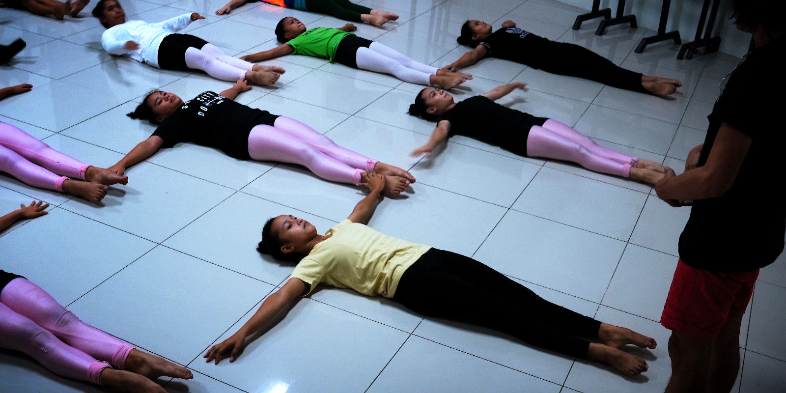 7 teenage female ballerinas lying on their backs on cold white floor, with arms stretched to the side and legs straight.