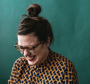 Photo of Daniella Sanader, laughing and looking down off camera. Her gold and teal patterned shirt contrasts vibrantly with the blue-green background.