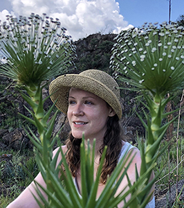 Headshot of Grace Grothaus outside on a summer day. There is a scrubby mountain in the background and two large spikey flowers in the foreground framing her face.