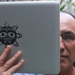 A headshot of Archer Pechawis with half his face obscured by a mac laptop with a Haida sun mask sticker over the apple