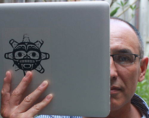 A headshot of Archer Pechawis with half his face obscured by a mac laptop with a Haida sun mask sticker over the apple