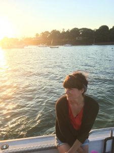 Mila Volpe in the foreground, on a boat at sunset. There is the lake and a small island in the middle ground.
