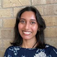 A portrait photo of Graduate Research Associate Sheetal Prasad wearing a dark blue shirt with white floral accents in front of a light yellow brick background.